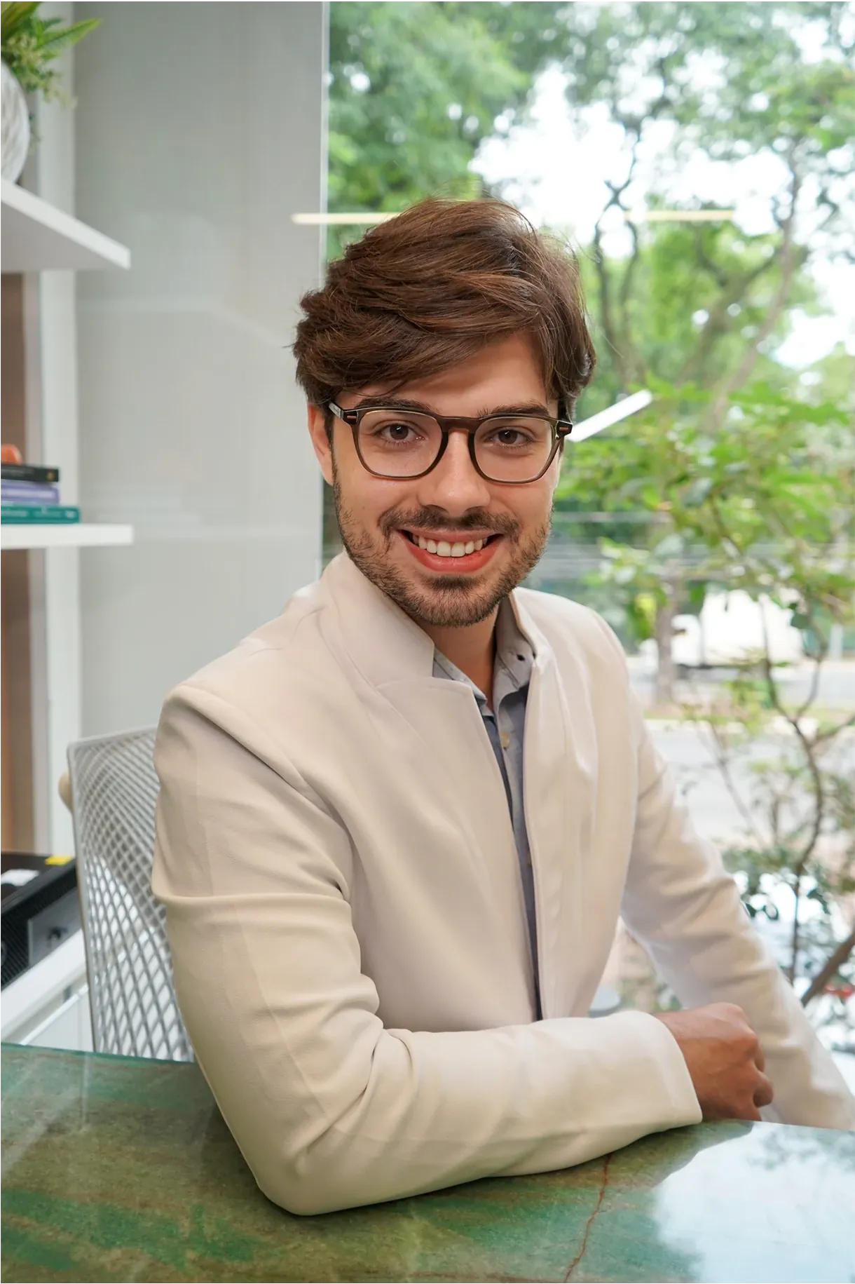 Dr. Caíque Dias in a light blazer, seated at a desk, smiling